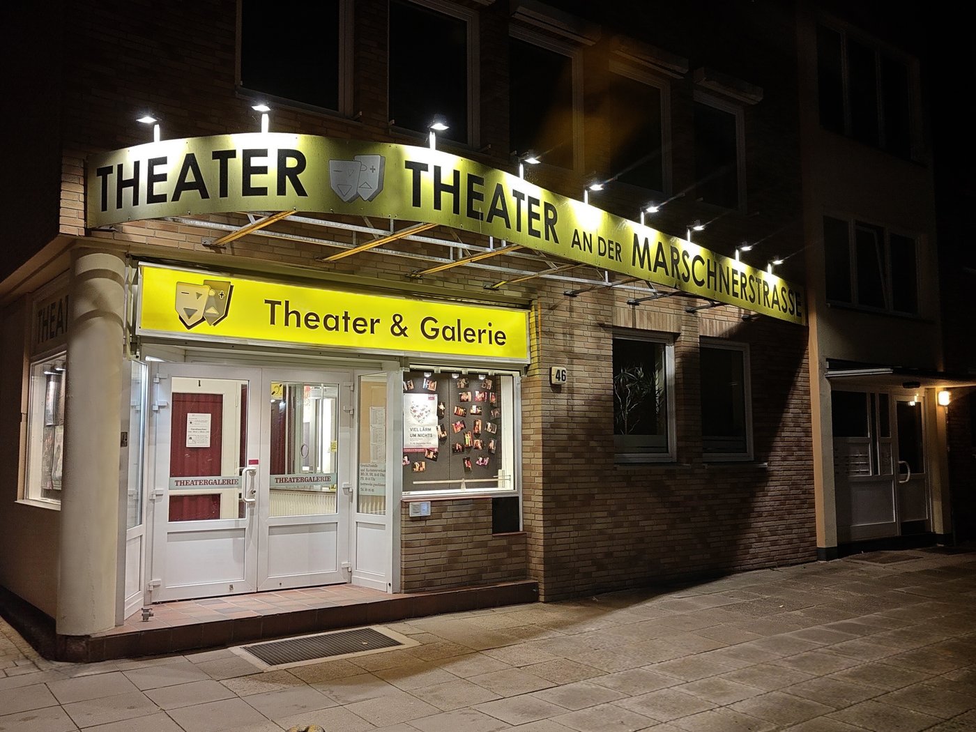 Das Theater an der Marschnerstrasse bei Nacht: beleuchtetes gelbes Schild „Theater an der Marschnerstrasse“ über der Ecke, darunter der Eingang mit dem Schild „Theater & Galerie“